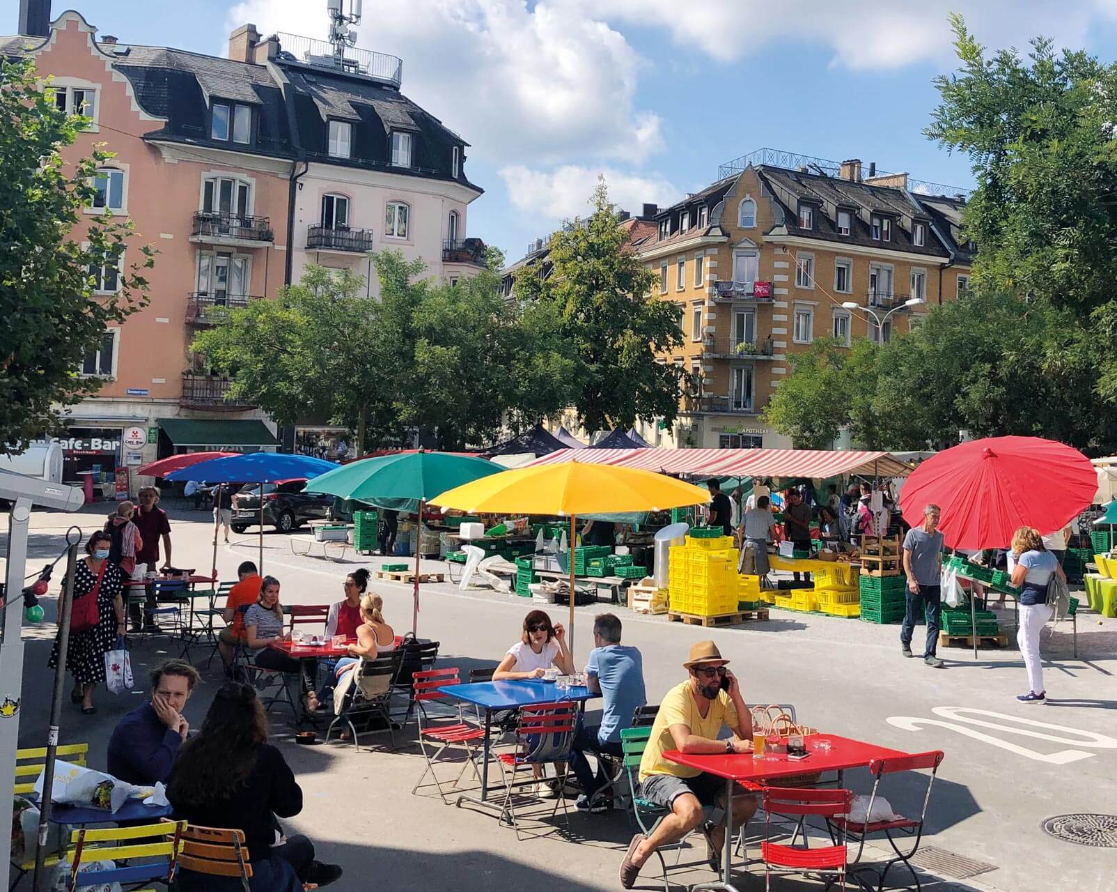Menschen sitzen unter Sonnenschirmen im Strassencafé. Nebenan ist der Wochenmarke auf dem Röschibachplatz.